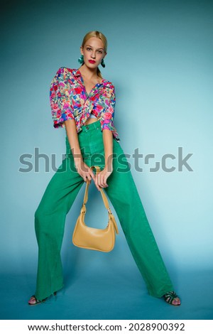 Summer fashion: confident blonde woman wearing colorful blouse, green high waist wide leg jeans, holding trendy yellow leather bag, posing on blue background. Full-length portrait Royalty-Free Stock Photo #2028900392