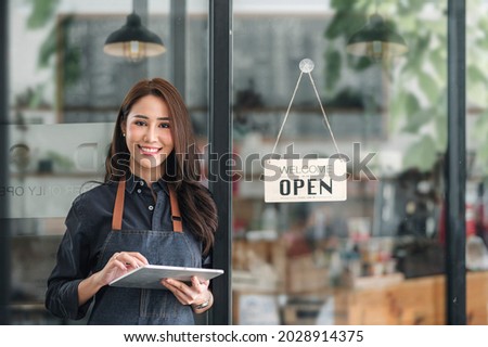 Beautiful asian young barista woman in apron holding tablet and standing in front of the door of cafe with open sign board. Business owner startup concept.
