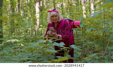 Senior elderly grandmother naturalist adventurer exploring forest trees, plants with mobile phone in wood. Tourist woman grandparent making photos using telephone. Active retirement, tourism, hobby