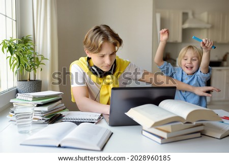 Teenager boy study at home and his younger brother screams and interrupts him. Online education and distance learning for high school students. School boy doing his homework using gadgets. Royalty-Free Stock Photo #2028960185