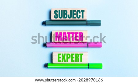 On a light blue background, there are three multi-colored felt-tip pens and wooden blocks with the SUBJECT MATTER EXPERT