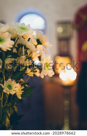 Bouquet of white chrysanthemums flowers on blurry background of church interior, with burning candles and icons near arched window