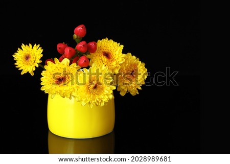 A small yellow mug with yellow chrysanthemums and hypericum on a glossy background. Close up, black background, negative space on the right. Still life, a beautiful autumn picture for a postcard.