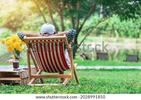 Back view of senior man with white hat sitting on garden chair and by the table in garden. Summer vacation in green surroundings. Happy person outdoors relaxing on deck chair in garden.Outdoor leisure
