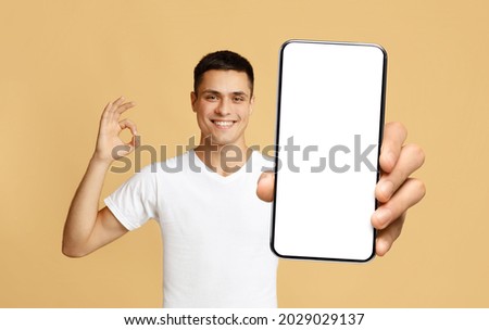 I Recommend. Portrait of smiling man holding smartphone with white blank device screen in hand, showing okay sign gesture, beige studio. Gadget with empty free space for mock up, isolated display Royalty-Free Stock Photo #2029029137