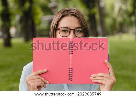 Young woman student freelancer teacher hiding her face with copybook notebook dairy relaxing sitting on green grass lawn doing homework after classes lectures at university college.