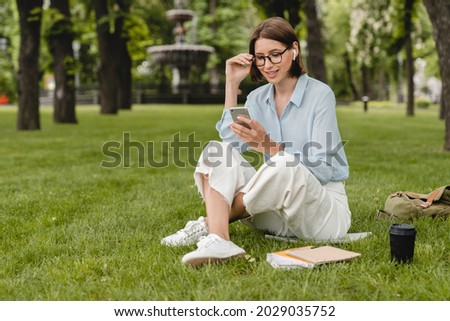 Caucasian young female student freelancer businesswoman relaxing on green grass lawn in city park after classes at university college, doing homework, surfing social media, texting, messaging online