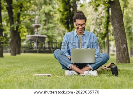 Young businessman student freelancer tutor using laptop for doing homework, e-learning, distant remote work job occupation, checking e-mails, e-banking online sitting on green grass lawn