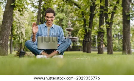 Young mixed-race student businessman talking having video call conversation online on laptop, vlogging blogging, watching webinar, lesson outdoors in city park on green grass lawn Royalty-Free Stock Photo #2029035776