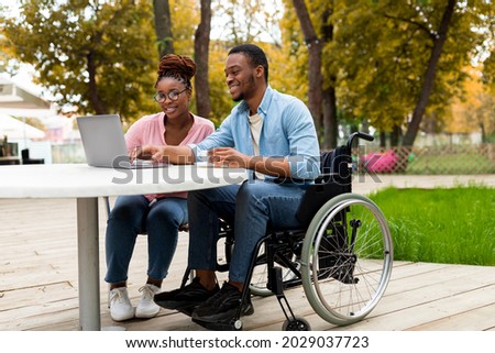 Cheerful impaired black man with female colleague using laptop together in outdoor cafe at autumn park, working on online business project, attending meeting remotely Royalty-Free Stock Photo #2029037723