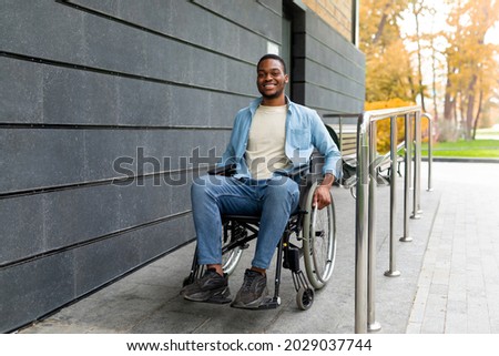 Handicapped accessible city concept. Positive impaired Afro man in wheelchair leaving building on ramp outdoors in autumn, full length. Joyful young black guy using disabled friendly facilities Royalty-Free Stock Photo #2029037744