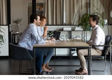 Smiling young Caucasian businesspeople talk with excited male candidate at job meeting in office. Happy man and woman recruiters speak with man applicant at interview. Recruitment, hr concept. Royalty-Free Stock Photo #2029043582