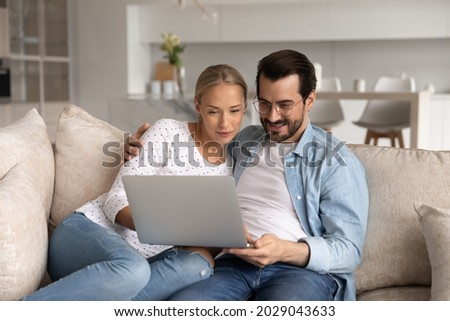 Happy millennial couple focused on laptop sitting together on sofa at home, hugging, using app on computer, watching movie online, shopping on internet, paying bills, insurance, mortgage fee Royalty-Free Stock Photo #2029043633