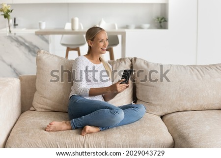 Happy mobile phone user resting on couch at home, looking away, thinking over good news. Blonde millennial woman using online app, virtual service on smartphone, texting, making call
