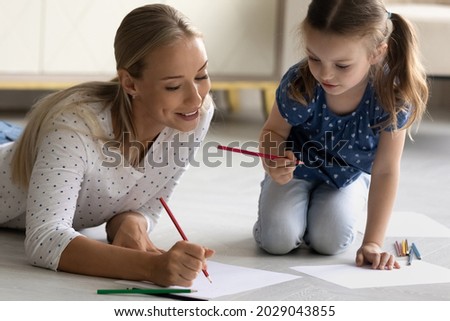 Happy mom teaching little daughter girl to draw with color pencils. Mum and kid relaxing on warm heating floor together, enjoying leisure time, home activities, developing creative artistic skills