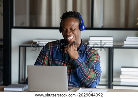 Overjoyed African American man in earphones sit at desk have webcam virtual event on laptop. Smiling young biracial male in headphones talk speak on video call on computer. Technology concept. Royalty-Free Stock Photo #2029044092