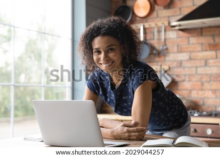 Happy African American student girl studying from home, standing by table with laptop, looking at camera with toothy smile. Female remote employee leaning at workplace. Head shot portrait