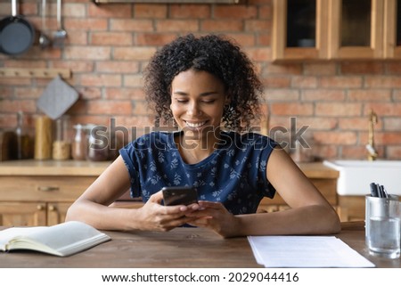 Happy Afro American student girl using smartphone at kitchen table, reading, texting, looking at screen. Black young woman, remote employee chatting online, checking messages, working from home