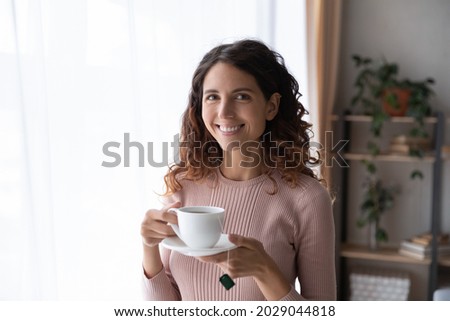 Happy young Latina woman standing in cozy room smile looks at camera holds cup of fresh brewed tea, enjoy no stress calm positive pastime, new day drink favourite beverage. Leisure, lifestyle concept