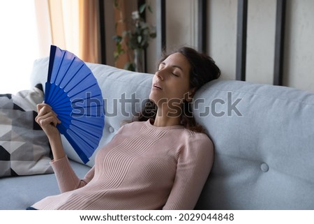 Overheated tired young Latina woman sit lean on sofa at home wave hand fan reduce heat, suffer from hot weather, lack of air conditioner indoor feels unwell exhausted, have hormonal imbalance concept Royalty-Free Stock Photo #2029044848
