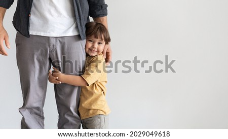 Father and son connection. Happy little boy embracing leg of his unrecognizable daddy, smiling to camera, feeling loved and safe, posing over grey wall background with empty space, panorama Royalty-Free Stock Photo #2029049618