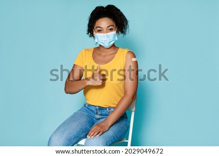 Portrait of young black woman with band aid after covid vaccination showing thumb up, approving of coronavirus immunization over blue studio background. Covid-19 vaccine saves lives concept