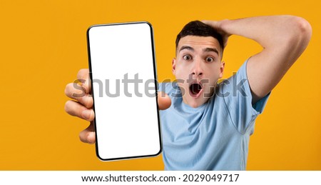 Wow, Super Huge Discount. Portrait of shocked guy grabbing head holding smartphone with white empty screen in hand, showing device close to camera, orange studio. Gadget display with copyspace, mockup