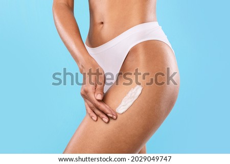 Treatment for Flawless Ideal Skin. Closeup cropped view of slim woman in white underwear panties applying body cream on hip isolated over blue studio background. Lady using moisturize lotion for legs