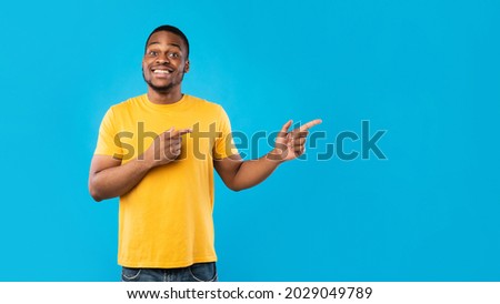 Look Aside. Black Male Pointing Fingers Showing Copy Space For Advertisement Text Standing Wearing Yellow T-Shirt On Blue Studio Background, Smiling To Camera. Panorama