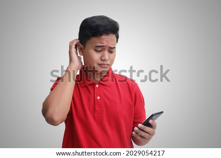 Portrait of unhappy Asian man in red polo shirt holding mobile phone with sad expression on face while scratching forehead. Isolated image on gray background Royalty-Free Stock Photo #2029054217