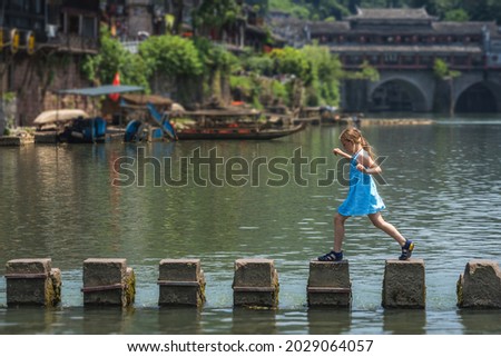 Young Caucasian girl crossing waters on stepping stones on Tuojiang river, flowing through the centre of Fenghuang Old Town, China Royalty-Free Stock Photo #2029064057