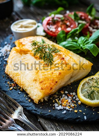 Fish dish - fried cod fillet with potatoes and vegetable salad served on stone plate on wooden table 