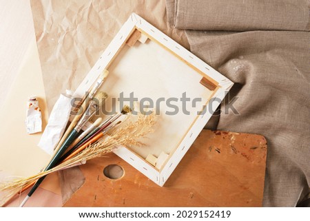 a set of brushes, paints in tubes, a wooden palette, a white canvas on a stretcher, paper and linen on a table, an artist's workshop concept top view