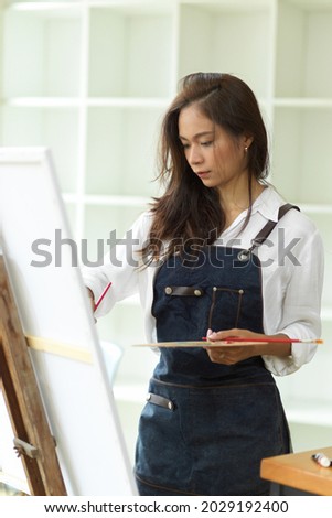 Attractive asian female artist working in her studio, passion painting, masterpiece, holding art palette, paint brush in hand