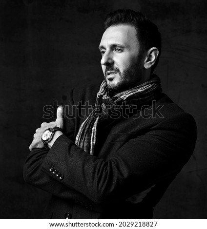 Black and white. Elegant bearded man in jacket and stylish scarf stands sideways, holding arms crossed at chest and looks right ahead over dark background