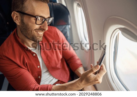 Cheerful caucasian man in casual wear and glasses traveling and taking picture of view outside through airplane window. Technology, travel, vacation, transportation concept
