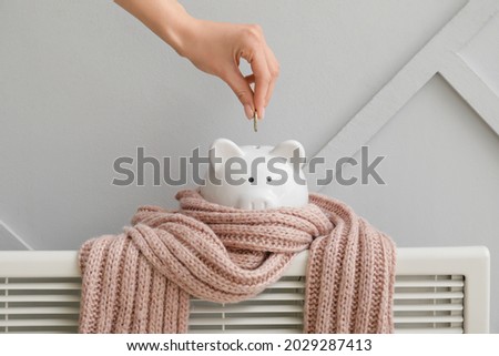 Woman putting money in piggy bank on radiator. Concept of heating season Royalty-Free Stock Photo #2029287413
