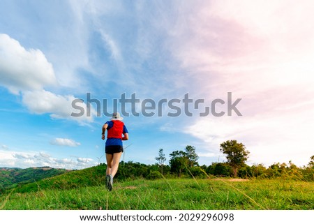 Behind the trail runner, an Asian woman wearing sportswear is practicing running on a high mountain behind a beautiful view. There is a field of wind turbines generating electricity in the background.