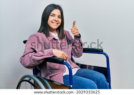 Young brunette woman sitting on wheelchair doing happy thumbs up gesture with hand. approving expression looking at the camera showing success. 