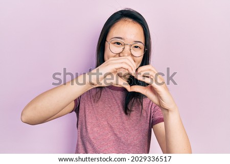 Young chinese girl wearing casual clothes and glasses smiling in love doing heart symbol shape with hands. romantic concept. 