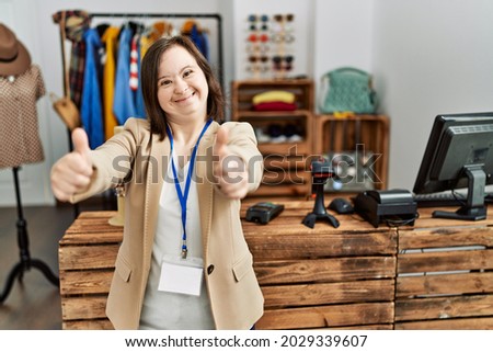 Young down syndrome woman working as manager at retail boutique approving doing positive gesture with hand, thumbs up smiling and happy for success. winner gesture.  Royalty-Free Stock Photo #2029339607