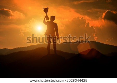 Silhouette of a businessman holding a trophy on top of a mountain with the sunset. The concept of a successful business or great executive to lead the organization to success. Royalty-Free Stock Photo #2029344428