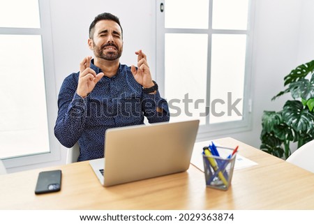 Young hispanic man with beard working at the office with laptop gesturing finger crossed smiling with hope and eyes closed. luck and superstitious concept. 