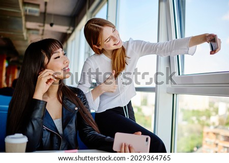 Cheerful Asian and Caucasian female bloggers smiling at front smartphone camera while shooting vlog content, happy multiractial hipster girls using mobile application for clicking selfie images