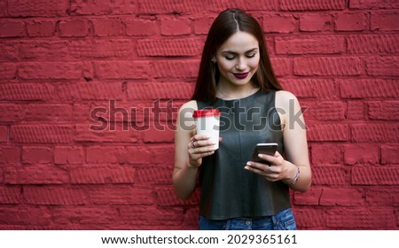 Millennial female blogger with coffee to go browsing network website for reading received email message, Caucasian hipster girl using mobile application for booking and banking near backdrop