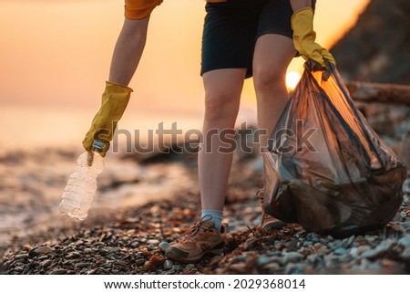 Earth day. Cleanup garbage on the ocean coast. A volunteer with polyethielene bag picking up a plastic bottle on the beach. Close-up. Sunset in the background. The concept of conservation of ecology. Royalty-Free Stock Photo #2029368014