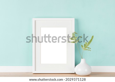 White vertical picture frame with matte and vase with fern leaves in front of pastel green wall. Elegant poster mockup. Blank image area for isolated with clipping path.