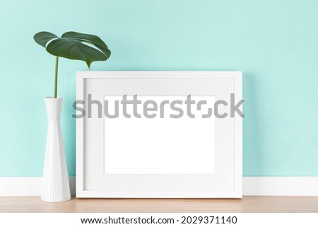 White picture frame with matte and monstera deliciosa leaf in vase in front of light turquoise wall. Elegant poster mockup, blank image area isolated with clipping path.