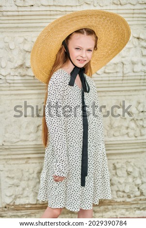 Elegant summer child girl in a wide-brimmed straw hat on a against the background of the textured wall. Kid's fashion. 