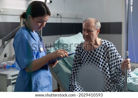 Senior sick man in hospital room discussing with nurse about treatment during consultation. Medical staff with stethoscope writing notes on clipboard preparing patient for surgery.